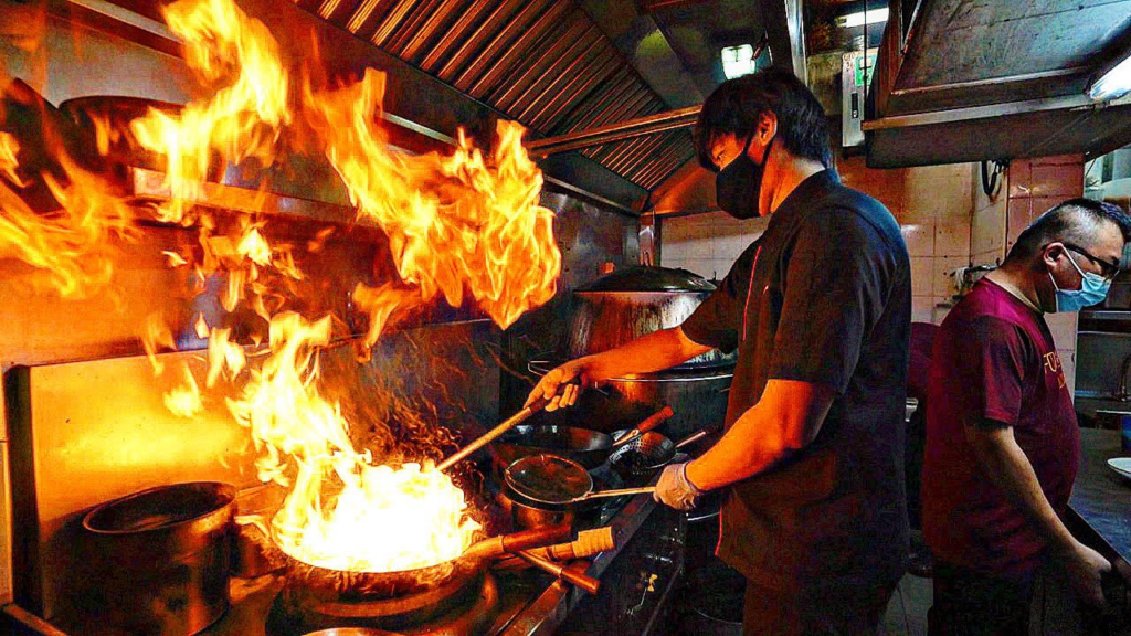 A chef preparing Char Kway Teow at a bustling Penang hawker stall, a key part of the Penang food experience. Tripmonks
