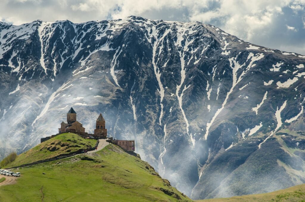 An ancient stone church sits on a green hilltop with a massive, snow-capped mountain rising in the background